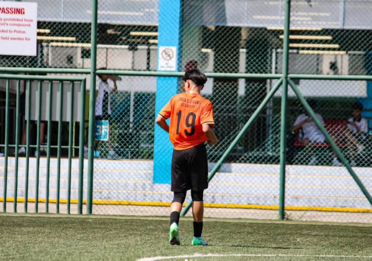 A soccer player in an orange jersey running on a green field during training near a building.